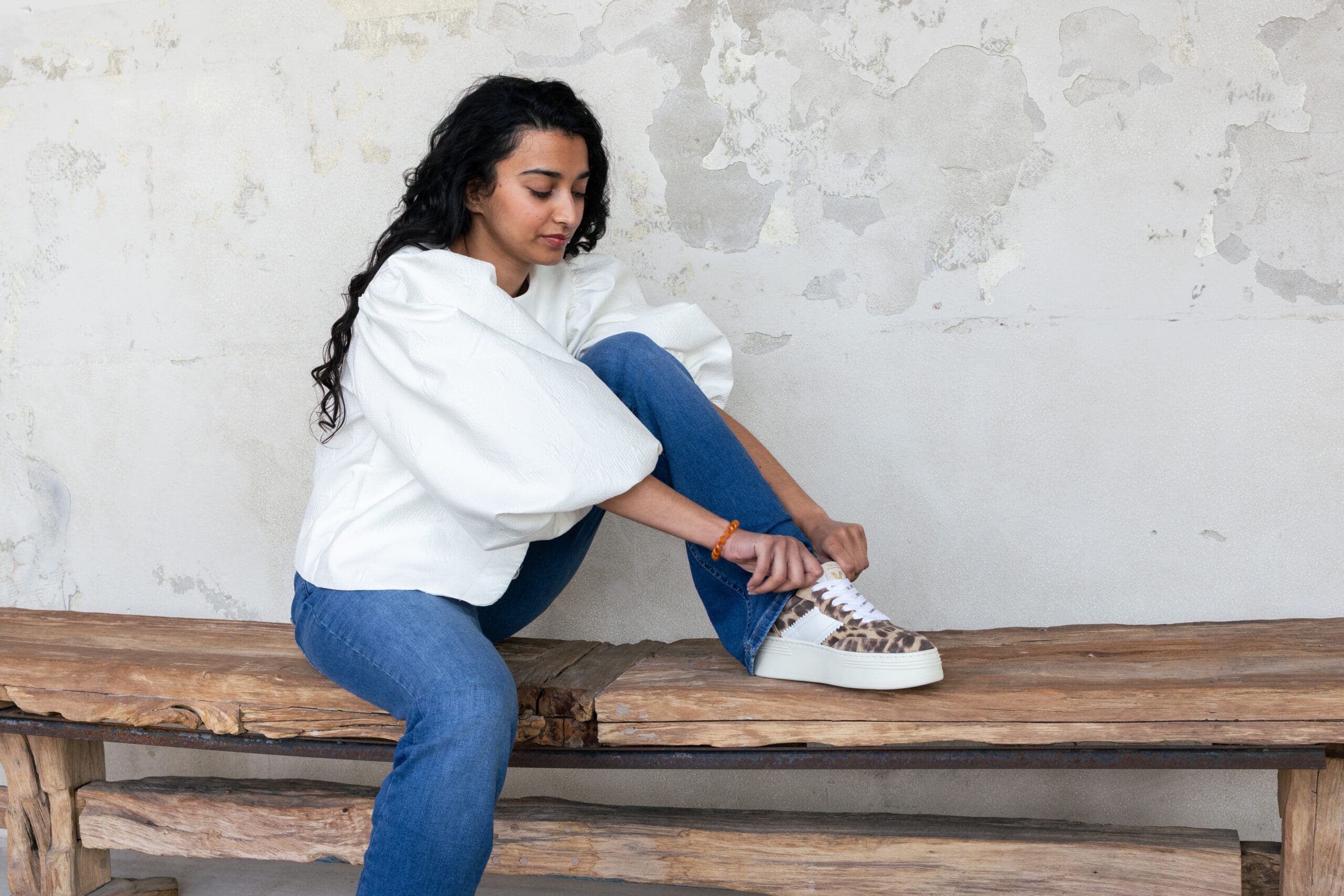 vrouw met witte blouse en spijkerbroek zit op bankje en strikt veter van haar panterprint sneaker - kledingtips voor brandingshoot